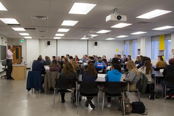 Students and teachers sit in a classroom listening to a speaker.