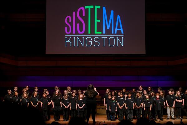 Sistema Kingston students stand on a stage wearing black. A large projector reads "Sistema Kingston" behind them.