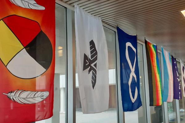 Flags hung inside a building including a flag with the medicine wheel, a flag with the Anishinaabe thunderbird, a flag with the Métis infinity symbol as well as a pride flag and trans pride flag.