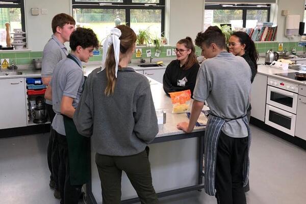 6 students and teacher candidates sitting around a kitchen bench creating a meal and laughing