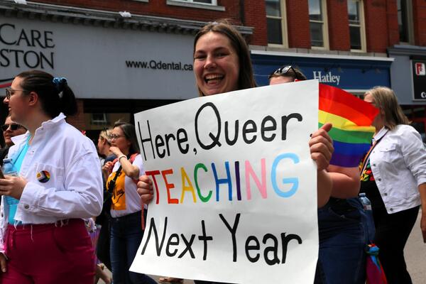A woman carries a banner that says, "Here, Queer, Teaching Next Year"