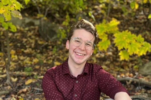 A white man with light hair sits on a rock with khaki pants on and a red shirt 