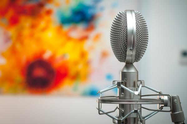 A silver microphone is in the foreground. The background has an out-of-focus colourful painting.