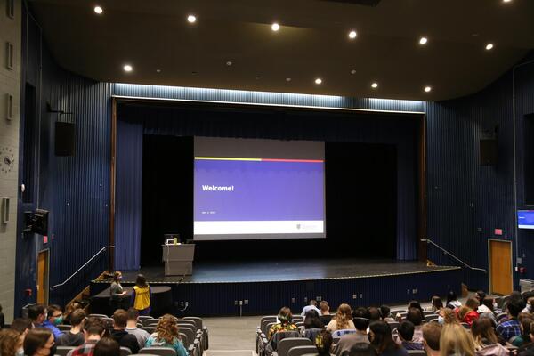 Students sit in an auditorium with a large screen at the front that reads "Welcome."