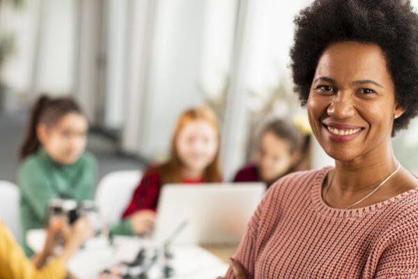 a teacher smiling at the camera with students working on a laptop in the background