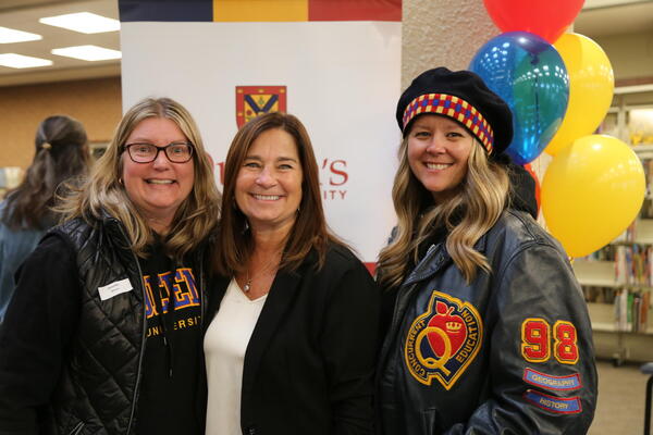 Three alumni stand in front of a banner and balloons. The woman on the right wears a tam and a Queen's leather jacket.
