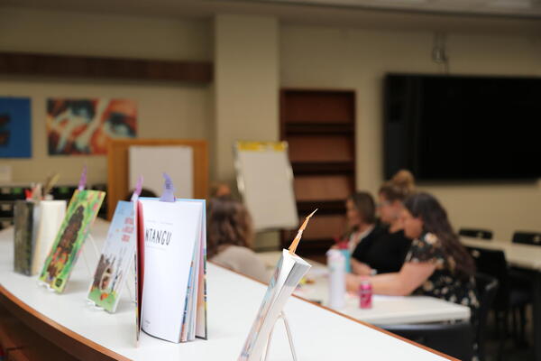 Books sit on a shelf in a library in front of a group of people working at a table.
