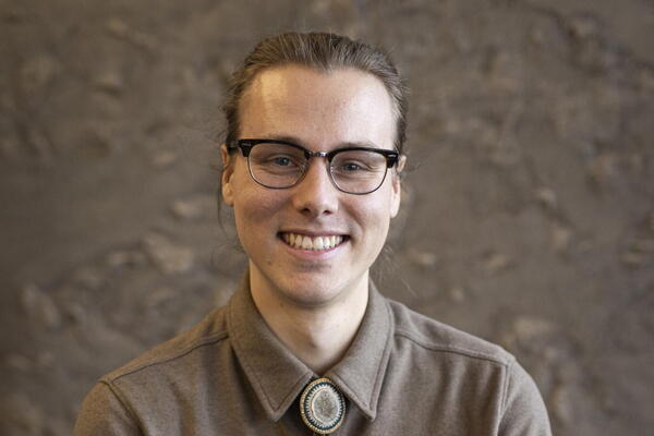 Jackson Pind smiles wearing glasses, a brown button down shirt, and a bolo tie in front of a mottled brown background.