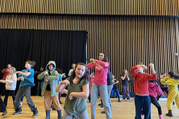 Olivia White leads Sistema students in a dance. Olivia wears a pink Sistema shirt and circles her arms in the air. The Sistema students have their arms in motion around Olivia.