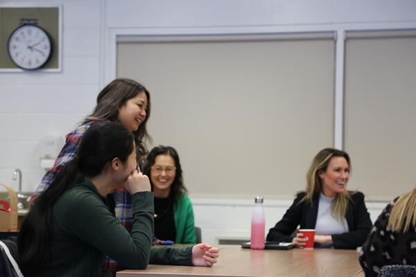 A group of students and professors laugh and smile around a table. There are water bottles and coffee cups on the table.
