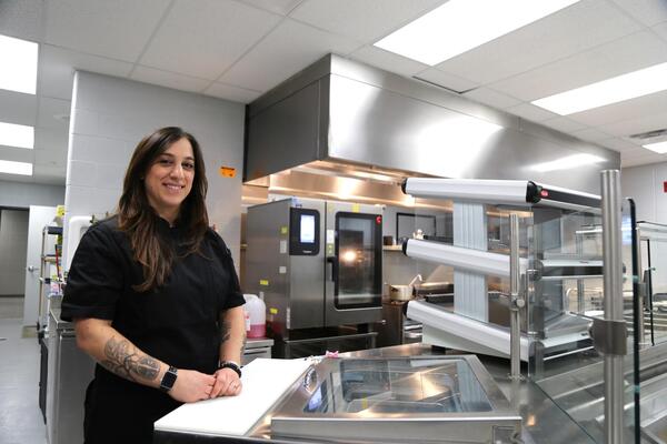 Grace smiles at the camera in an industrial kitchen that is full of stainless steel appliances and surfaces