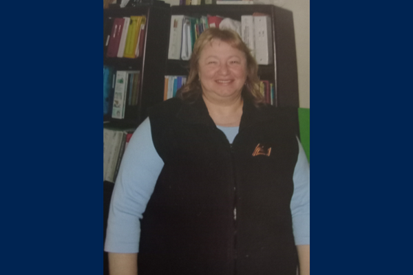 Connie Rosario smiles wearing a vest and blue shirt and standing in front of a bookshelf.