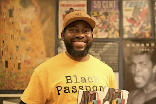 Paul Akpomuje smiles holding two copies of his book. Paul wears a yellow t-shirt with black writing that reads "Black Passport."