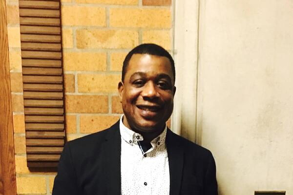 Leroy Baker smiles wearing a collared shirt and blazer and stands in front of a brick wall.