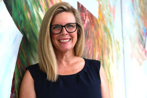 A woman smiles at the camera with blond hair and wearing glasses and posing in front of art 