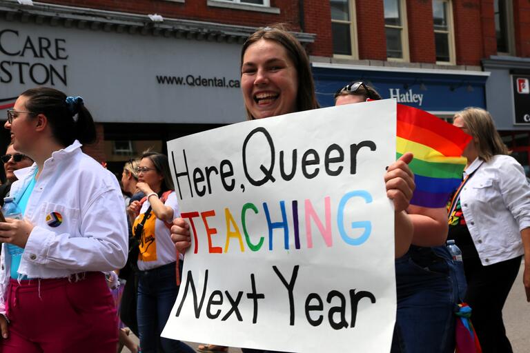 Smiling student carrying a sign that reads "Here, Queer, Teaching Next Year."