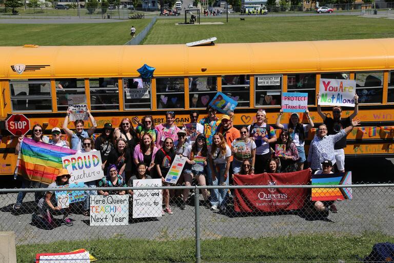A group of students wearing rainbow outfits in front of a bus holding signs that say things like "Here and teaching next year"