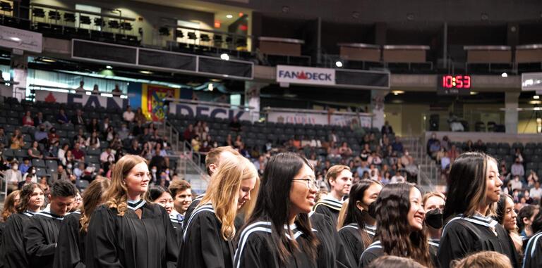 Students in graduation gowns face a stage with spectators observing.