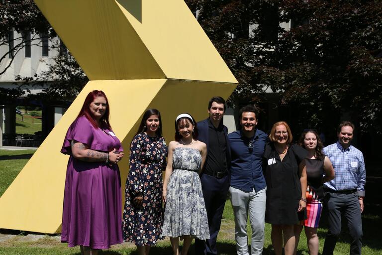 Graduates stand smiling in front of a large yellow outdoor sculpture.
