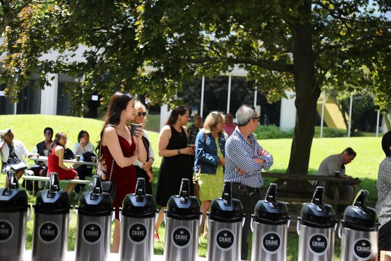 A graduate stands listening to a speech holding a coffee cup.