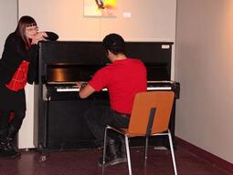 A teacher candidate playing piano and another teacher candidate leaning on the piano listening.
