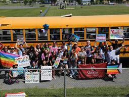 A group of students wearing rainbow outfits in front of a bus holding signs that say things like "Here and teaching next year"