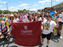 Student and Faculty Members holding up a banner that says Queen's Faculty of Education while wearing vibrant rainbow colors outside