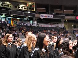 Students in graduation gowns face a stage with spectators observing.
