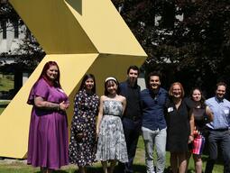 Graduates stand smiling in front of a large yellow outdoor sculpture.