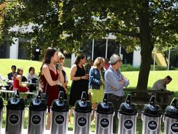 A graduate stands listening to a speech holding a coffee cup.