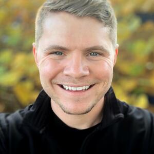 A white man smiling with blue eyes and blond hair with green outdoor space blurred in the background and a black shirt on