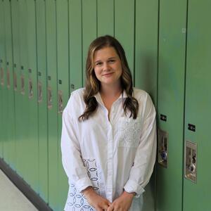 Vicky smiles at the camera. She has long light brown hair. She is wearing a white collared shirt and is leaning against a row of green lockers