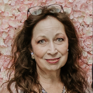 Aynne looks at the camera. She has wavy long dark brown hair. She is wearing a large necklace and earrings. Her glasses are sitting on top of her head.