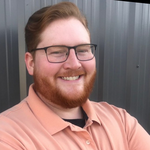 Bruce smiles at the camera. He is wearing a peach polo shirt and glasses. His short hair and beard are red.