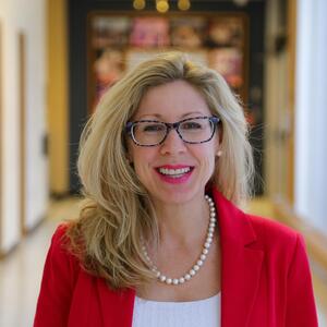Christine smiles at the camera. She is standing in a hallway wearing a bright red blazer and white blouse. She has long blond straight hair and has glasses and a pearl necklace.
