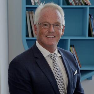 Terry smiles at the camera. He has short white hair and is wearing glasses and a blue suit jacket, white collared shirt and blue and white tie.