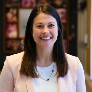 Erin is smiling at the camera. She is wearing a blush pink blazer, a white shirt and a necklace. She has dark brown straight hair that is longer than shoulder length.