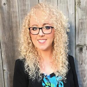 Winter smiles at the camera. She is wearing black glasses. She has curly long light blond hair. She is wearing a black blazer and a blue floral shirt.