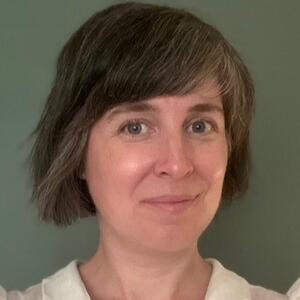 Holly grins at the camera. She is wearing a white blouse with a peter pan collar and has chin length light brown hair.