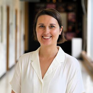 Nicole is standing in a hall smiling at the camera. She is wearing a white short-sleeved collared shirt and has chin length dark brown hair.