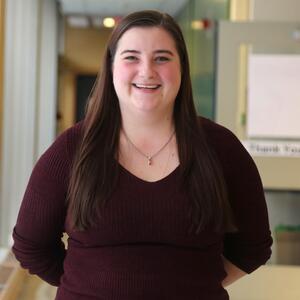Andrea is smiling at the camera. She has long dark brown hair and is wearing a necklace and dark purple shirt.