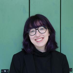 Belinda smiles at the camera. She has dark purple, shoulder length hair and is wearing a black shirt and round dark rimmed glasses.