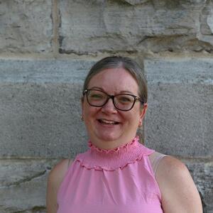 Lori smiles at the camera. She is wearing a pink sleeveless blouse and dark rimmed glasses. Her hair is pulled back.