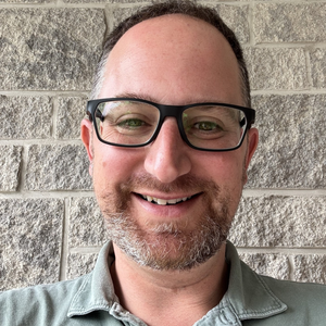 Jeff smiles at the camera. He has short brown hair and a short beard. He is wearing glasses.