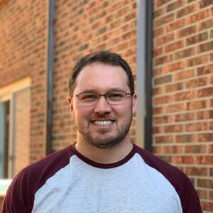 Josh smiles at the camera. He has short brown hair and is wearing glasses and a grey and maroon baseball t-shirt
