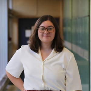 Kali grins at the camera. She has shoulder length straight, brow hair. She is wearing a white collared shirt and is wearing glasses.