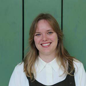 Mari smiles at the camera. She has long, straight blond hair and is wearing a white collared shirt and a dark green dress.  She is standing in front of some green lockers.