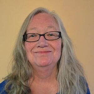 Pam smiles at the camera. She has long white hair and is wearing dark rimmed oval glasses and a blue shirt.