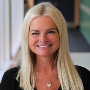 A woman with blond hair a gold necklace and black shirt smiles at the camera