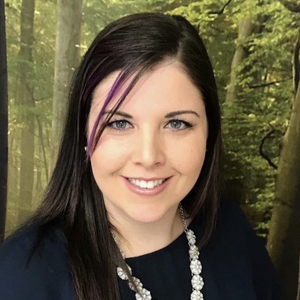 Heidi smiles at the camera. She has long dark brown hair with a purple streak. She is wearing a black shirt with a silver flower necklace.